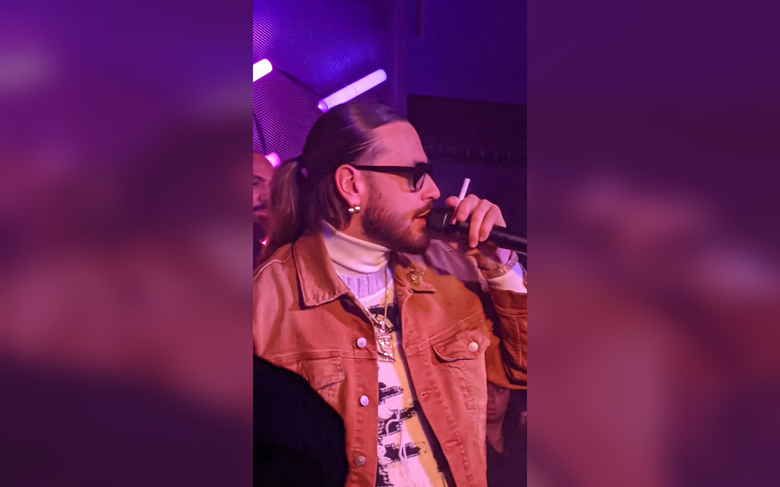 Rapper in sunglasses and orange denim jacket singing into a microphone onstage